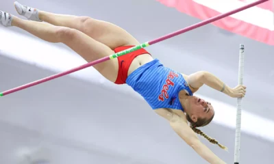 Ole Miss pole vaulter clears the bar during an indoor meet, fully extended mid-air in a blue Rebels uniform with an American flag blurred behind.