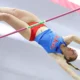 Ole Miss pole vaulter clears the bar during an indoor meet, fully extended mid-air in a blue Rebels uniform with an American flag blurred behind.
